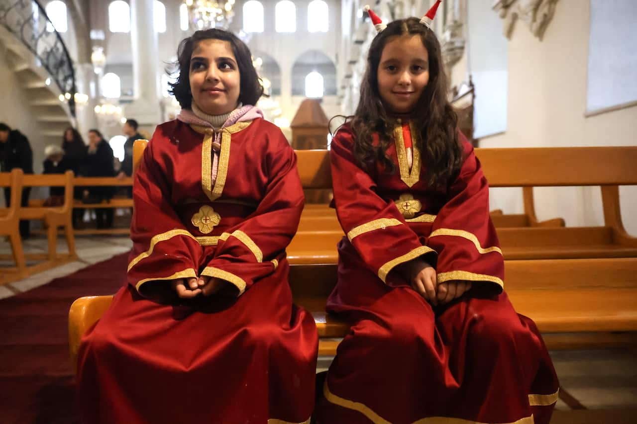 Two young girls in red dresses with golden borders on the tops sit on a pew inside a church.