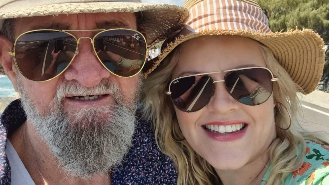 an older man in a sun hat, sunglasses and a grey beard smiles in front of a beach with a younger blonde woman who wears a sun hat and sunglasses