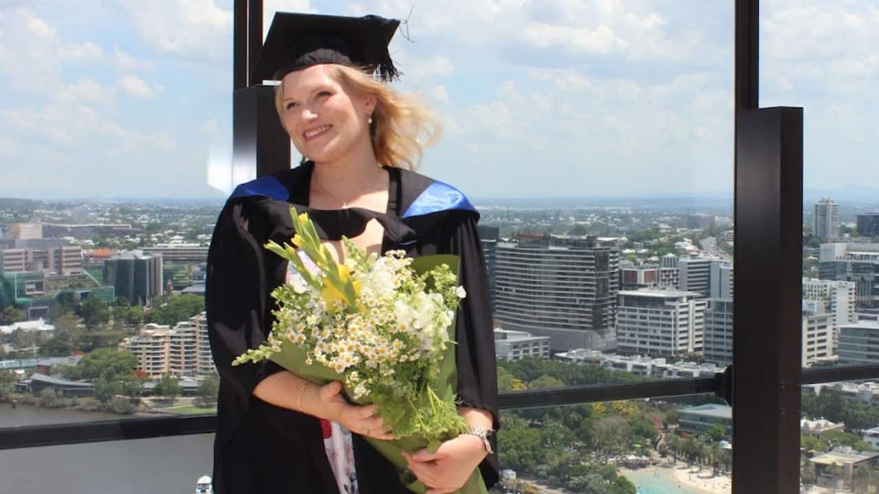 a young blonde woman in a graduation gown and cap holds a bouquet of flowers on a high rise balcony
