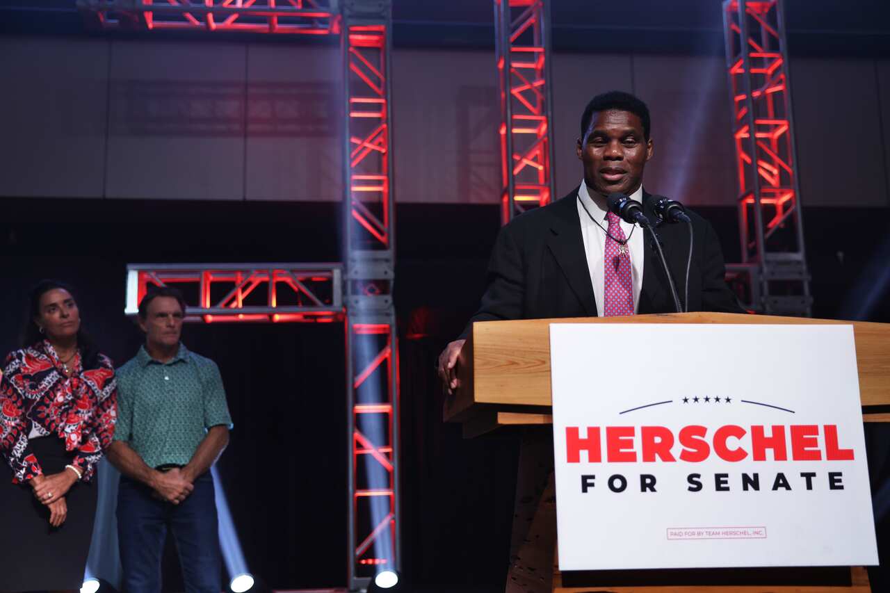 A man speaking at a podium with a sign attached that reads: "Herschel for Senate". 