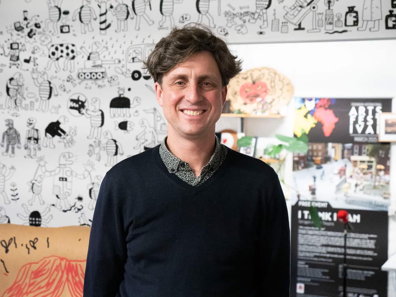 A man smiling at the camera. He is wearing a collared shirt and a black jumper 