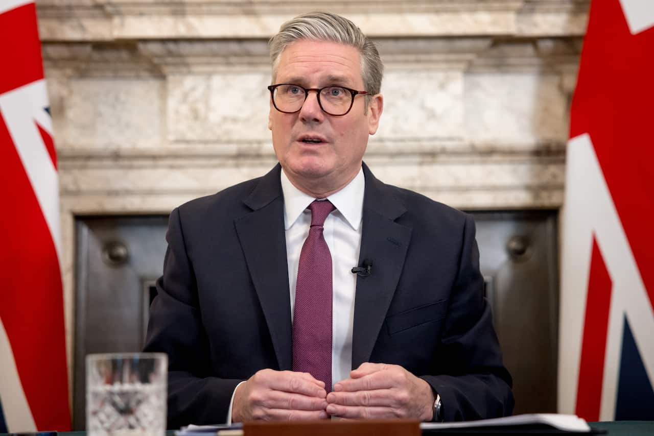 Keir Starmer wearing a suit, sitting at a table with Union Jack flags in the background.