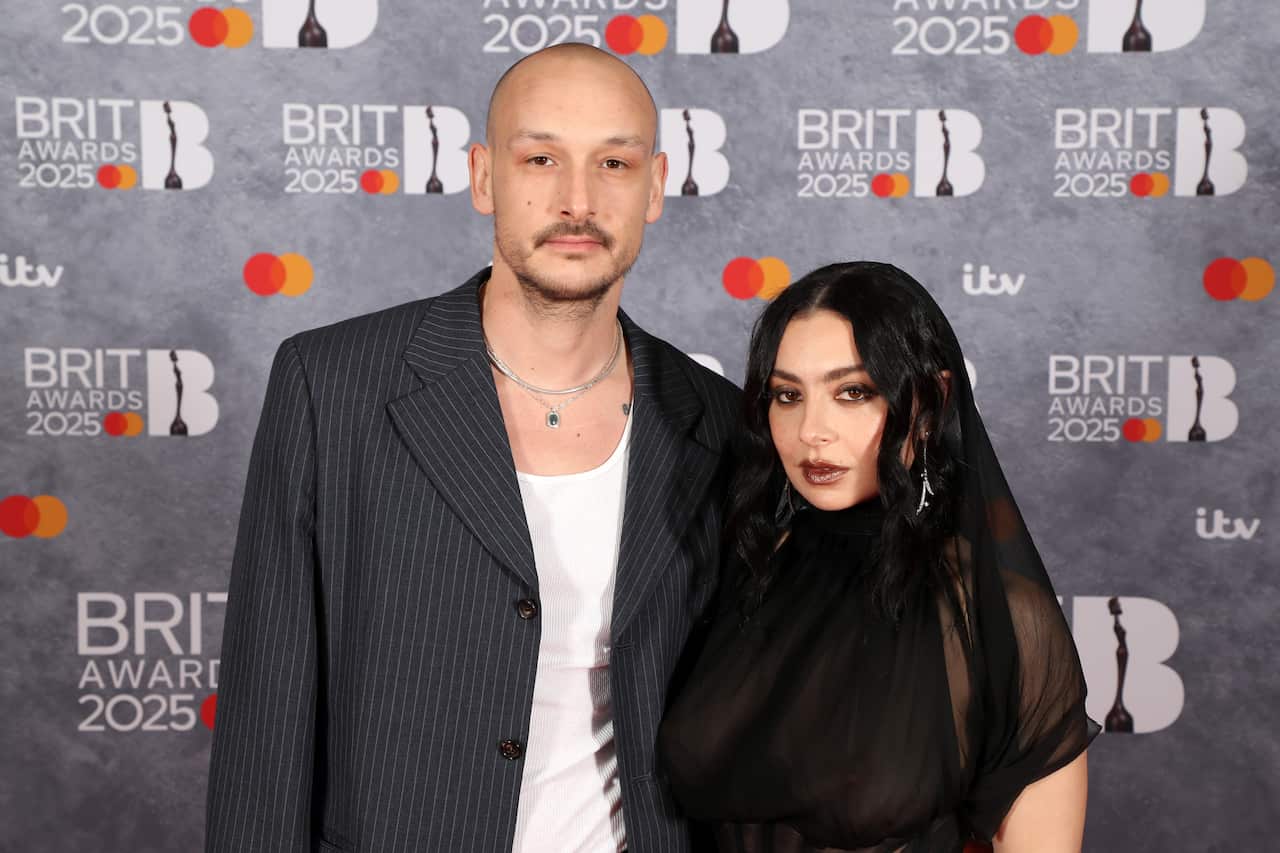 Charli XCX posing with her fiancé George Daniel. Behind them is a grey media wall with the 2025 Brit awards logo on it