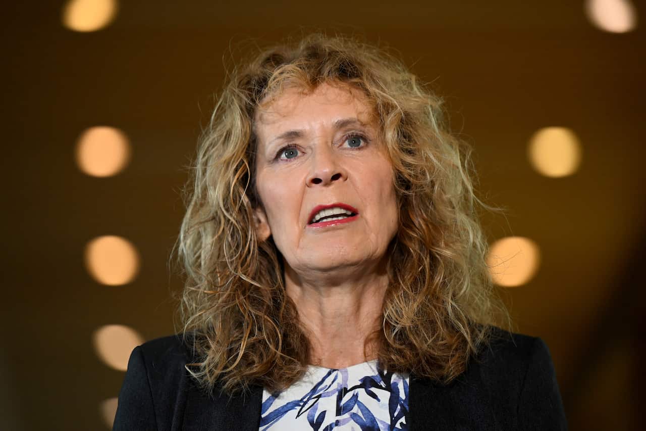 A woman with blonde wavy hair, wearing a navy blazer and white and blue top speaking