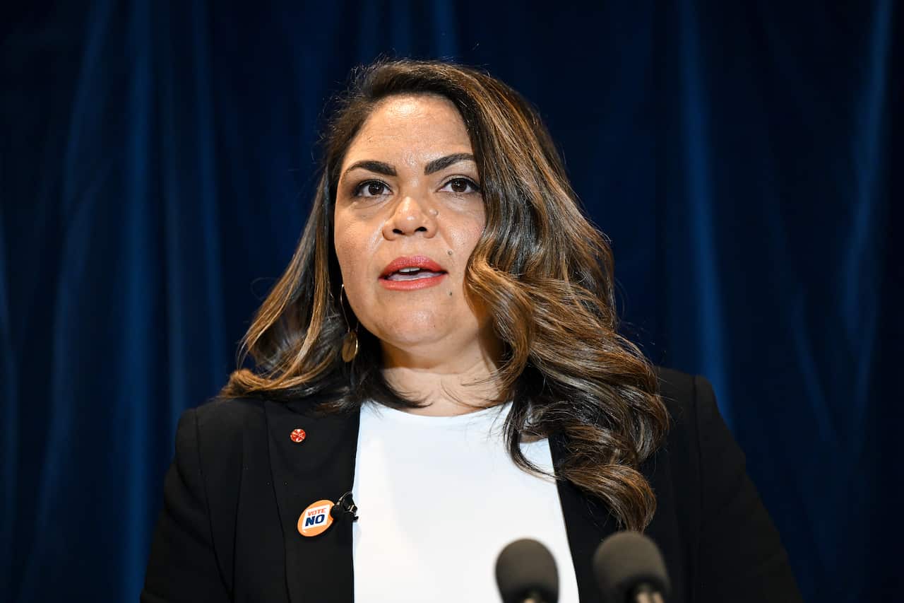 Jacinta Price stands in front of a microphone wearing a white shirt and black jacket.