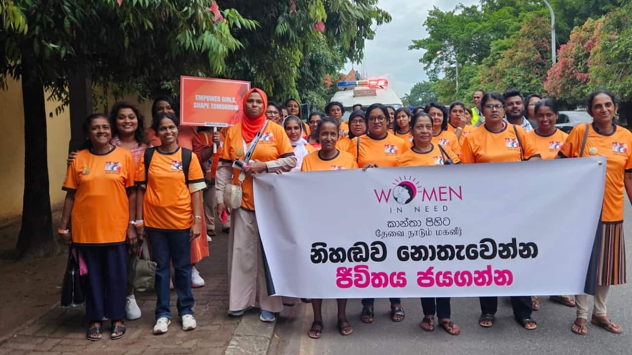 A large group of women wearing orange t-shirts poses with a banner with text in Sinhala language and the words Women In Need" in English at the top.