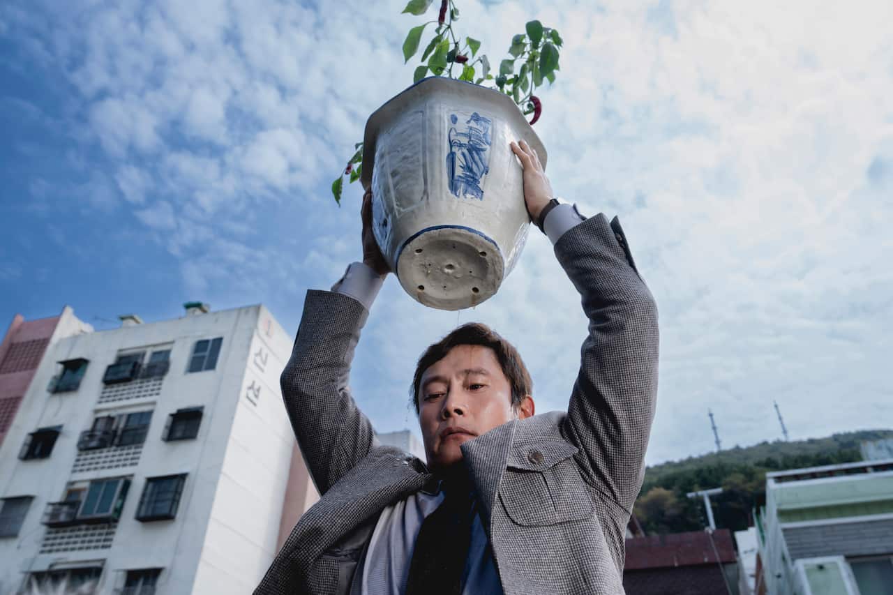 A man in a suit holding a vase. A blue sky is behind him. 