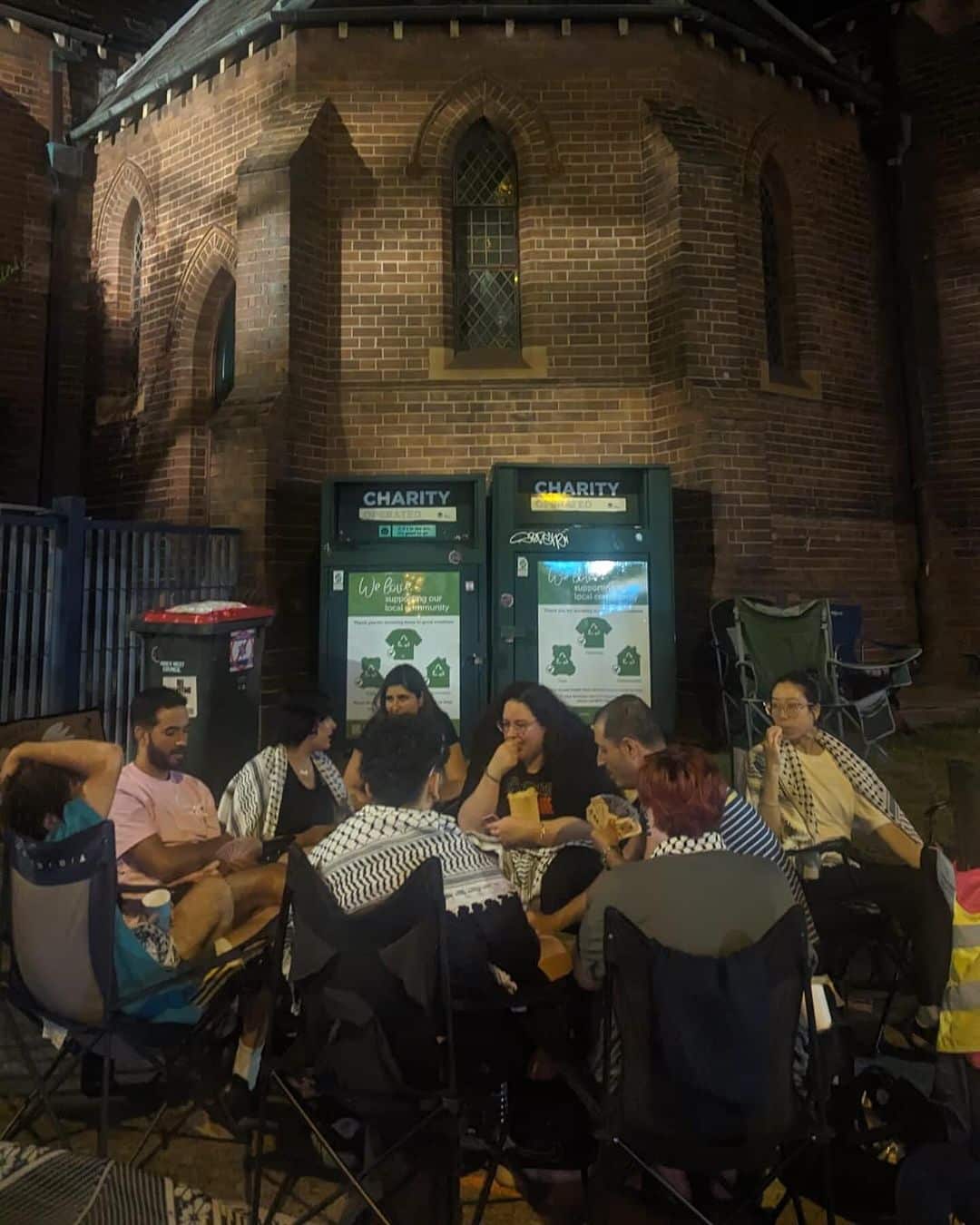 A group of people wearing Palestinian keffiyeh sit in a circle in front of the brick facade of a church
