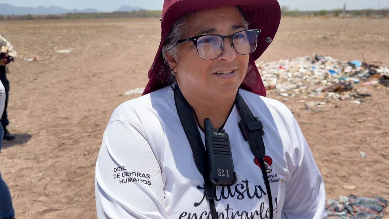 A middle-aged woman wearing glasses, a wide-brimmed burgundy sun hat and a white search group shirt stands in a dry, open field