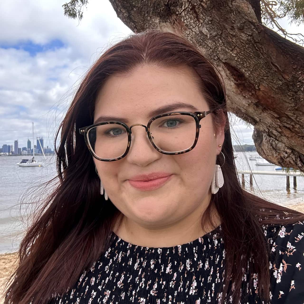 A woman with dark hair and glasses standing on a beach with city buildings in the background