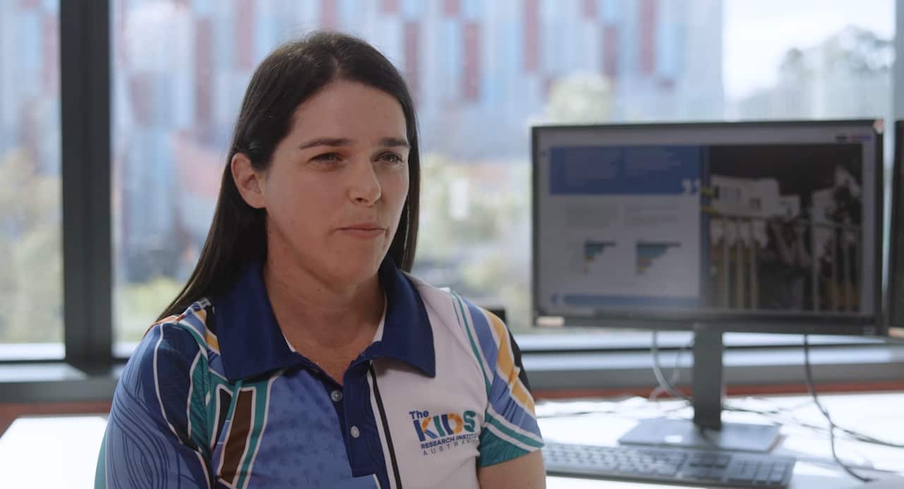 A woman wearing a blue polo shirt that reads "The Kids Research Institute" is sitting in an office. There is a computer screen behind her.