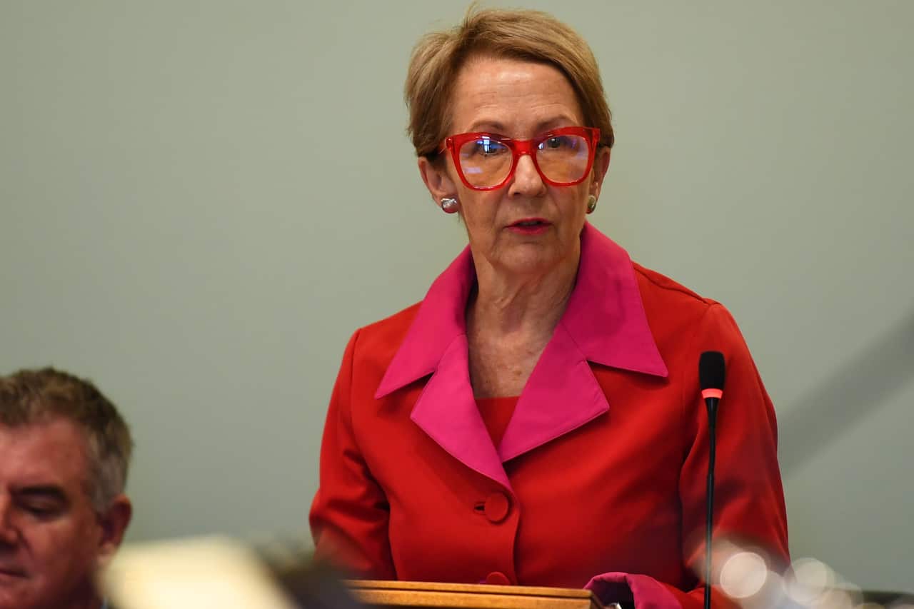 A woman with short, blonde hair wearing red glasses and a bright red jacket with pink lapels stands at a lectern.