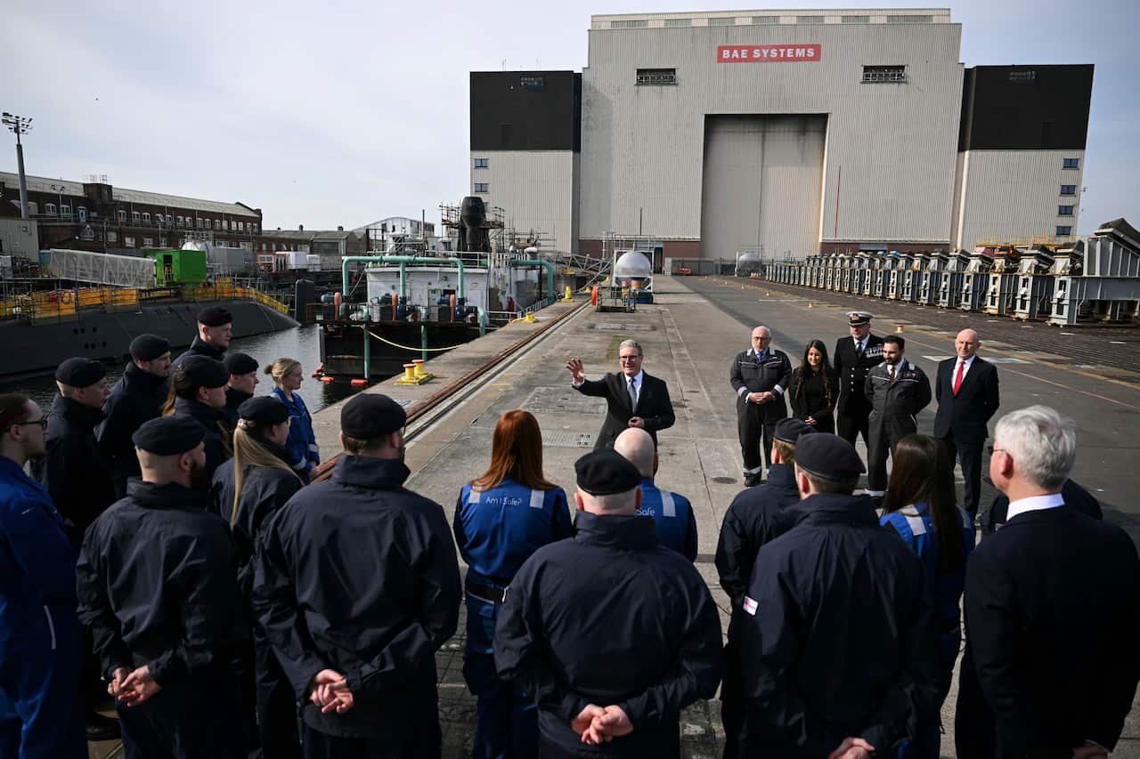 A crowd of military officers, sailors, politicians and submarine technicians gather at a port. A man wearing a suit stands in the middle of the crowd with his hand raised.