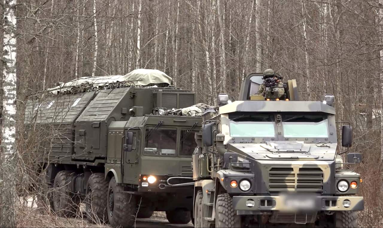 Two Russian military vehicles drive down a dirt path within a dense, leafless forest.