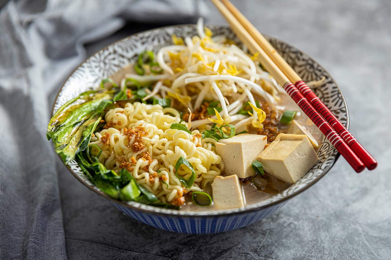 Chopsticks rest across one side of a bowl. The bowl has liquid at the bottom, topped with noodes, cubes of tofu and other toppings. 