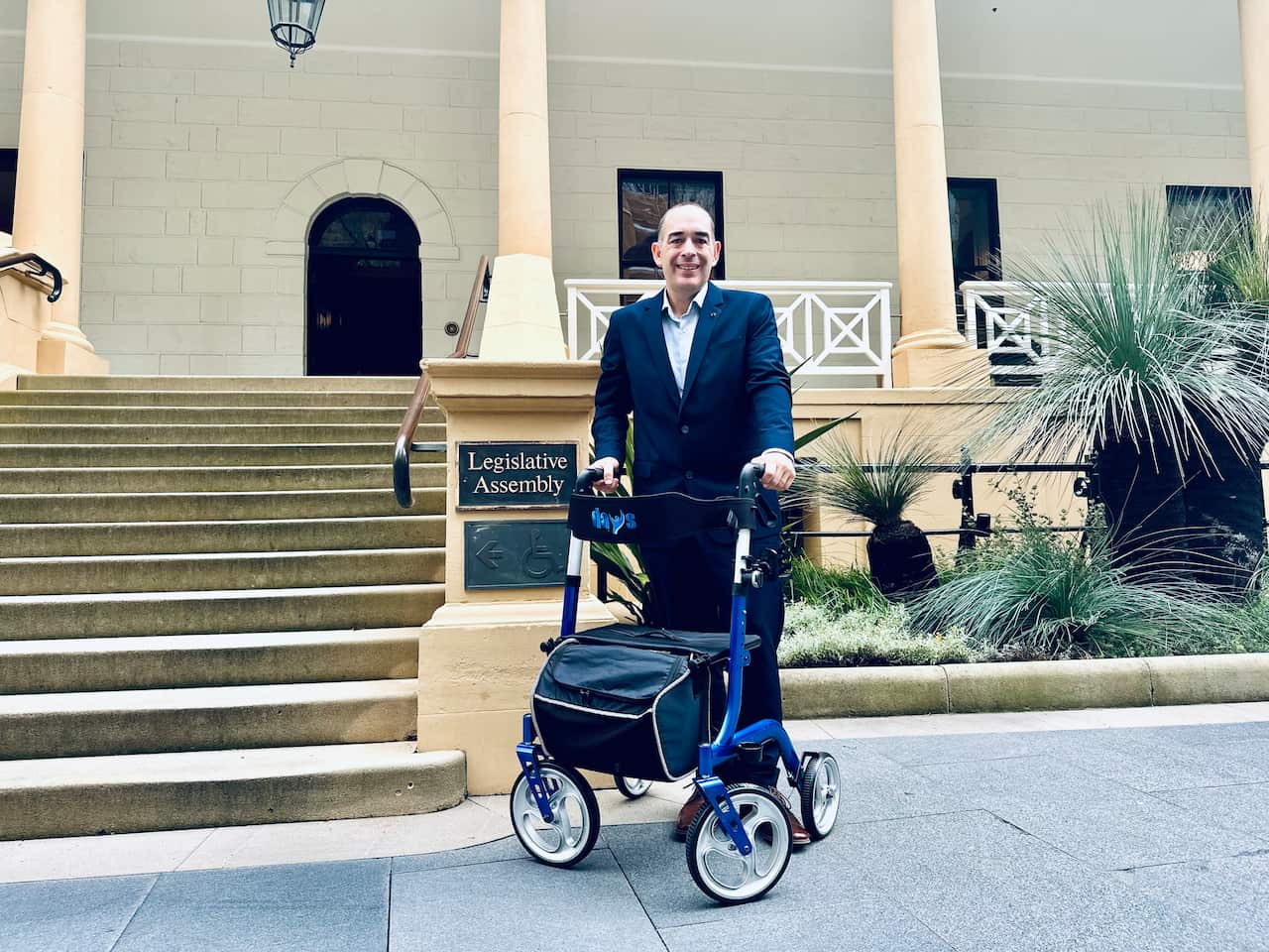 A smiling man in a suit stands on a paved area outside a government building, holding onto a blue four-wheeled walker. The building is beige with large columns and a flight of stairs leading to the entrance.