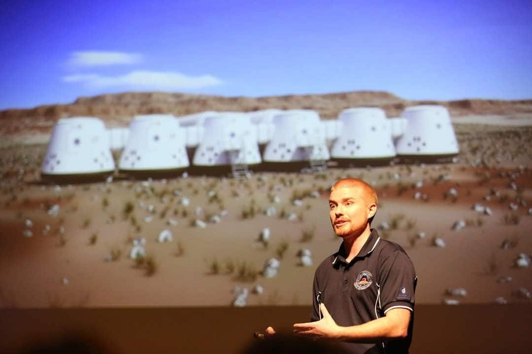 A man is delivering a talk with the backdrop of Mars one concept images on a large screen