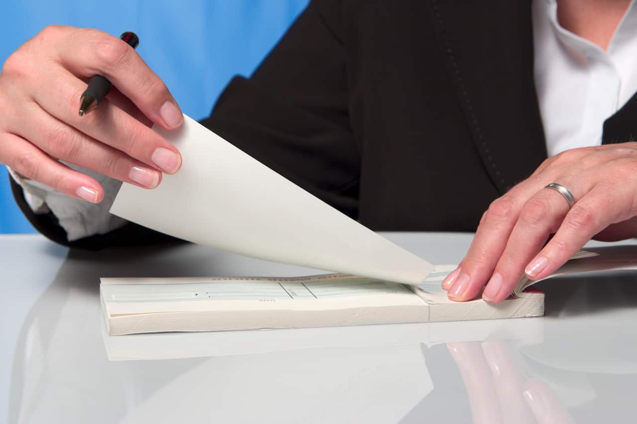A person's hand holding a chequebook, with a pen in their other hand ripping out a cheque.
