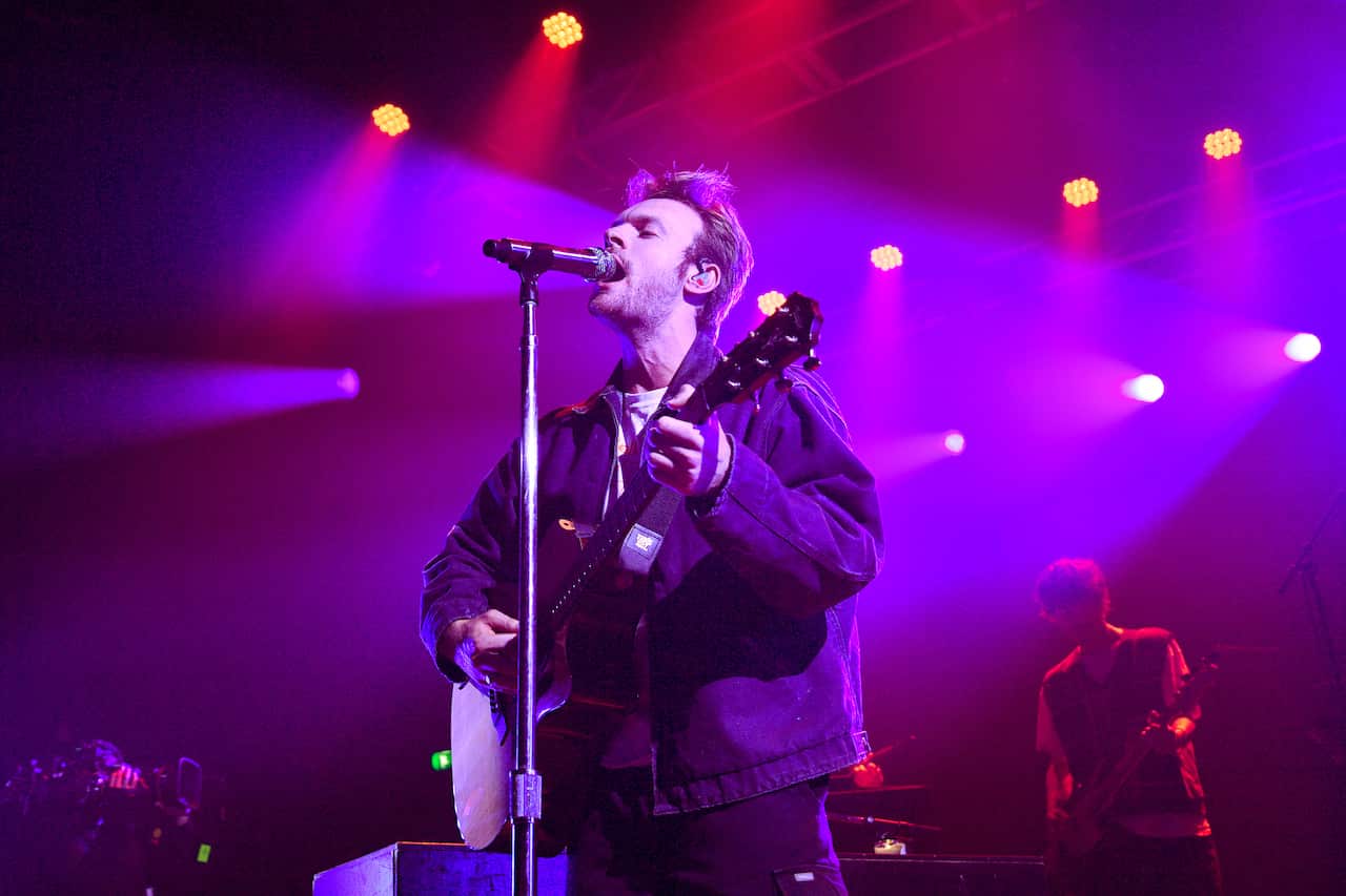 Finneas, wearing a dark parka, plays an acoustic guitar and sings into a microphone on a stand