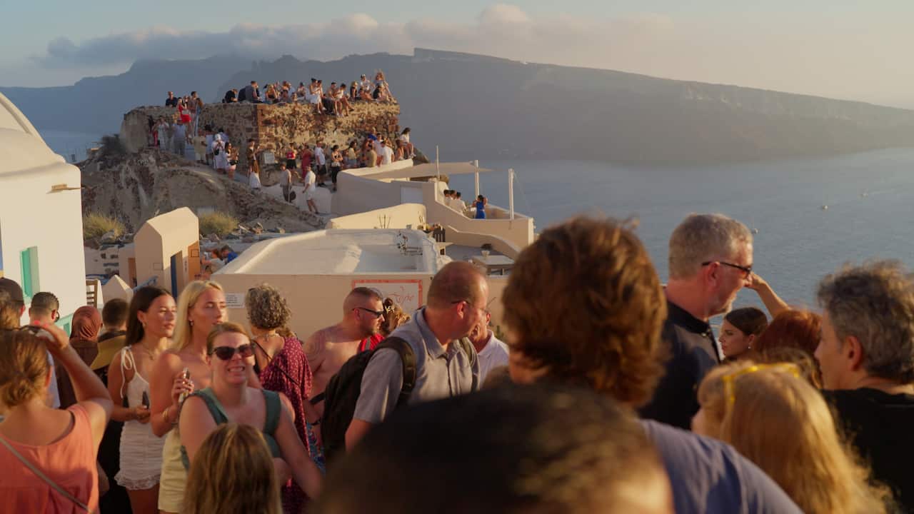 Crowds of tourists stand along rocky ledges and among white buildings as the sun sets over the ocean.
