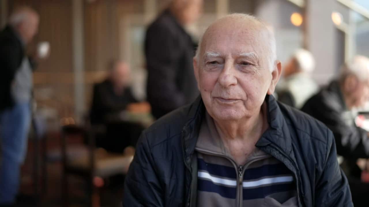 Older man with white hair sitting facing the camera, wearing a black jacket and brown and white striped inner jumper. Our of focus behind him are other old men sitting and standing around.
