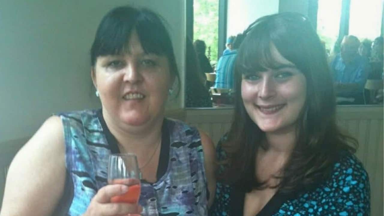 A middle aged woman with dark hair and a fringe smiles holding a glass next to a young woman with long brown hair smiling