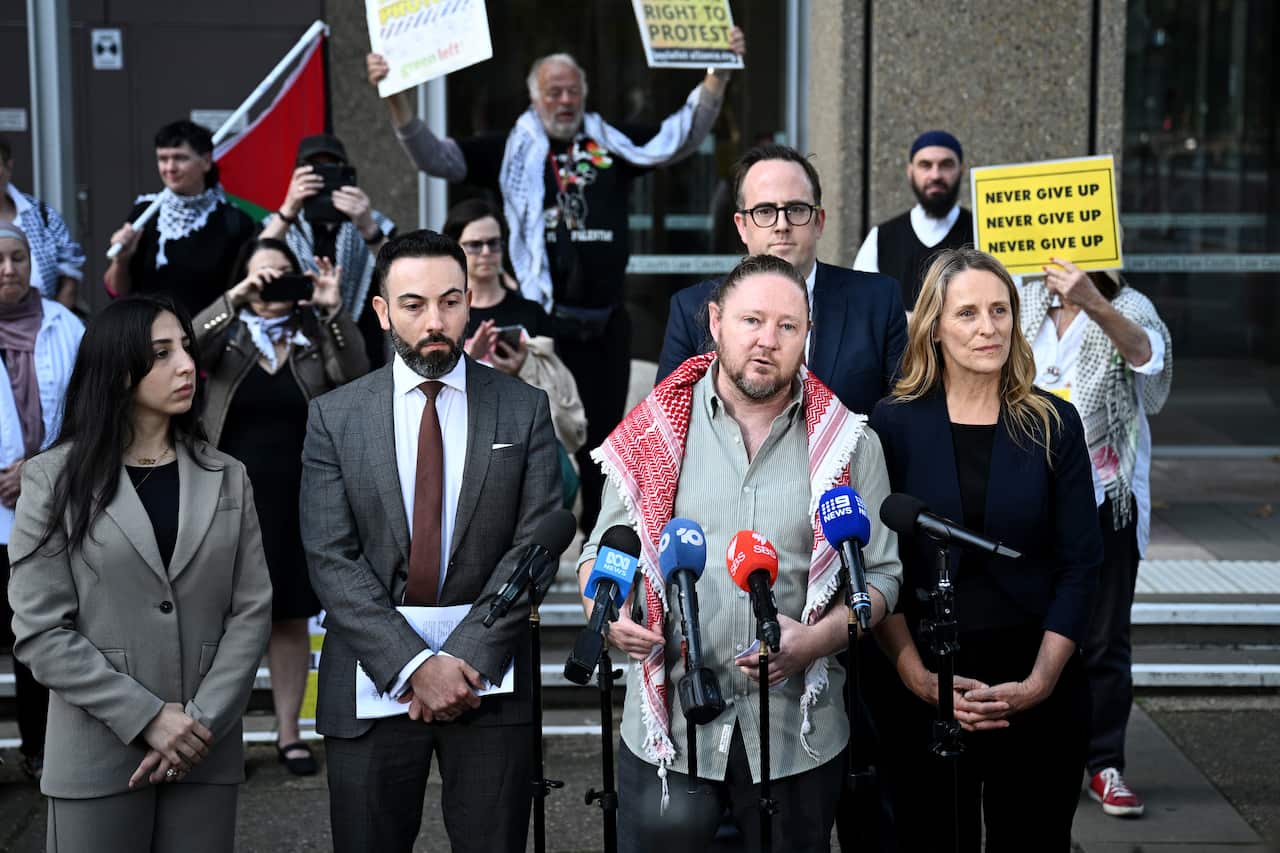 A group of activists and legal representatives, including a man wearing a keffiyeh around his shoulders, speak at a press conference held outside a courthouse while protesters hold signs and a Palestinian flag in the background.
