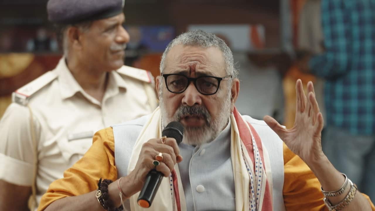 An elderly Indian man with grey beard and hair and wearing reading glasses and a traditional men's outfit is speaking into a microphone