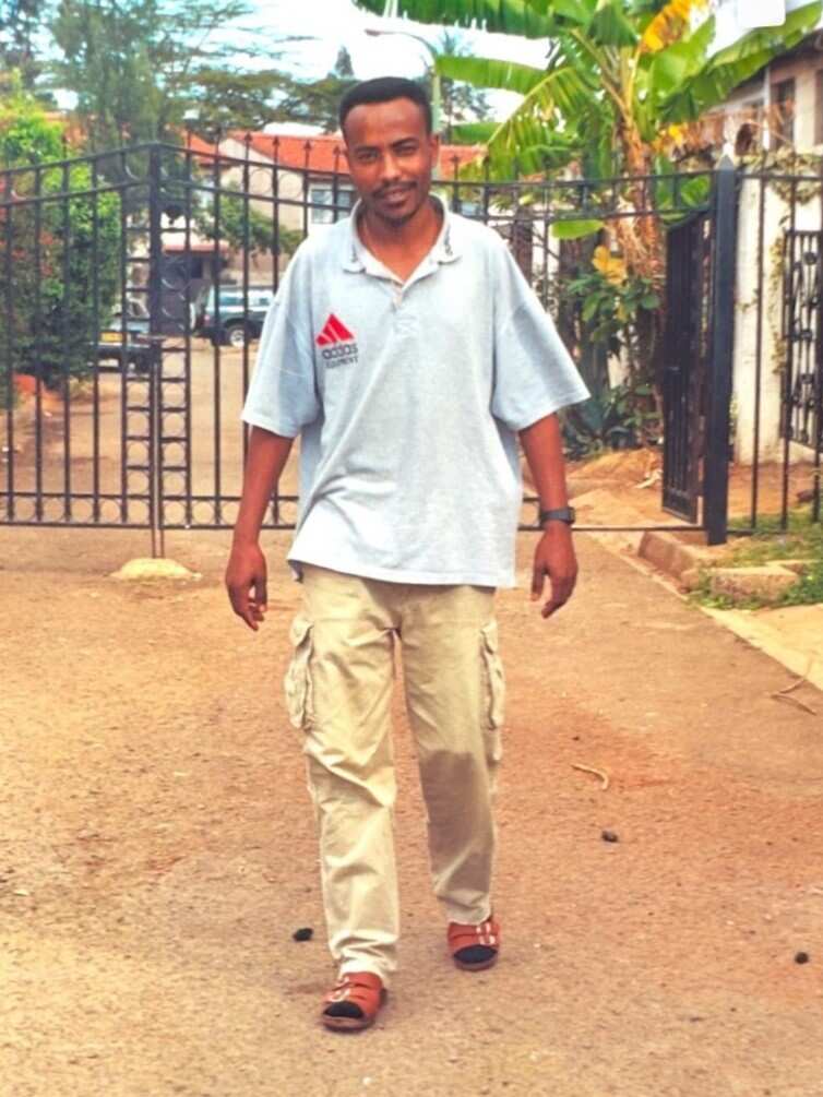 A young man in a light blue T-shirt stands in front of metal gates on a dirt path.