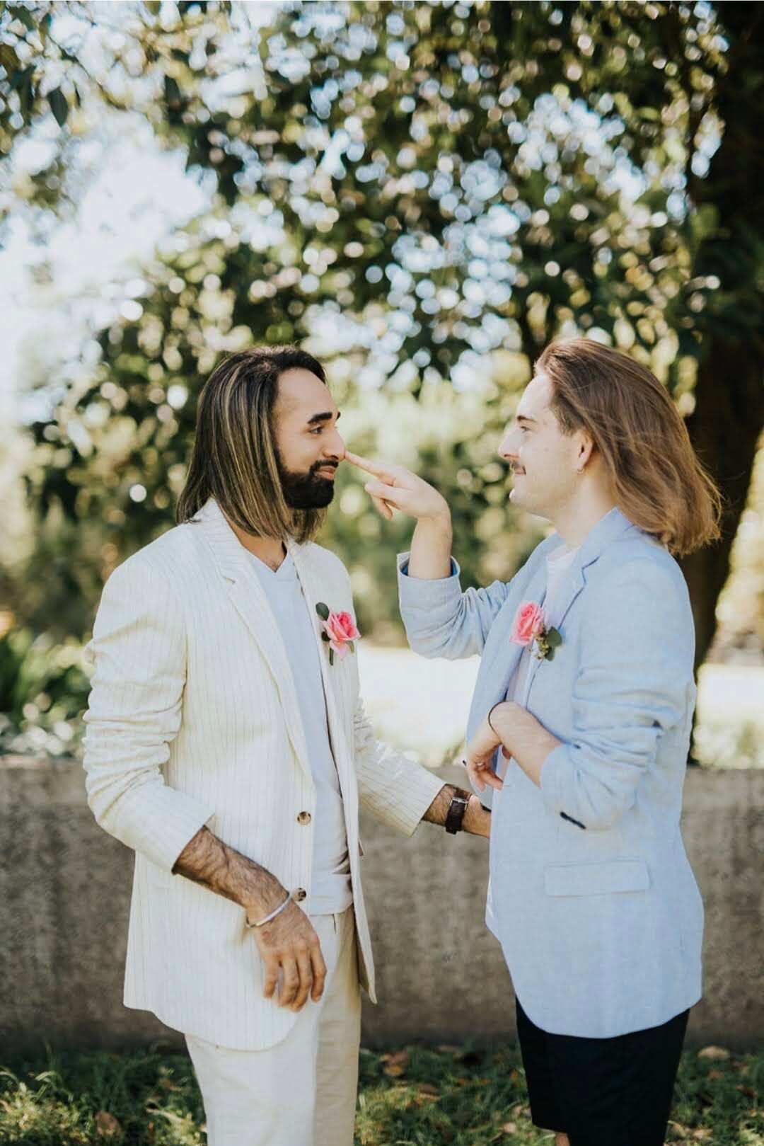Two gay men facing each other on their wedding day. The man on the left is wearing a white suit and the man on the right a light blue suit jacket and navy pants. They smile at each other as one lovingly touches the other's nose.