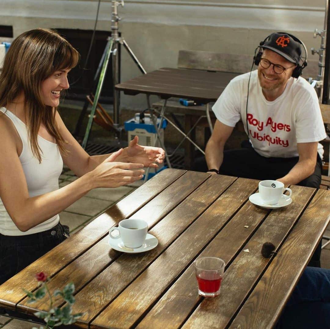 A woman gestures animatedly while talking to a smiling man wearing headphones and a "Roy Ayers Ubiquity" T-shirt at an outdoor cafe table with two mugs sitting on the table in front of them.