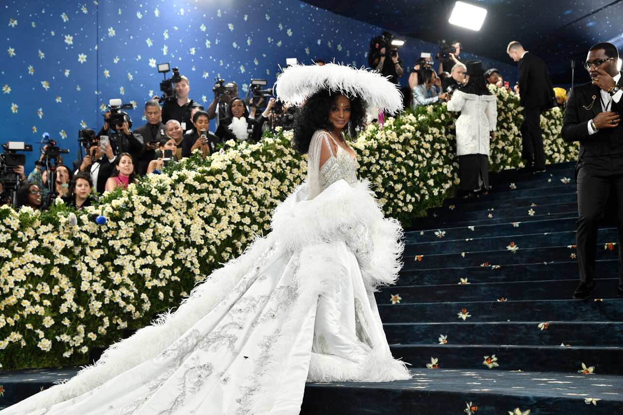 Diana Ross standing on stairs and wearing a white dress with a long train and white feather wide hat