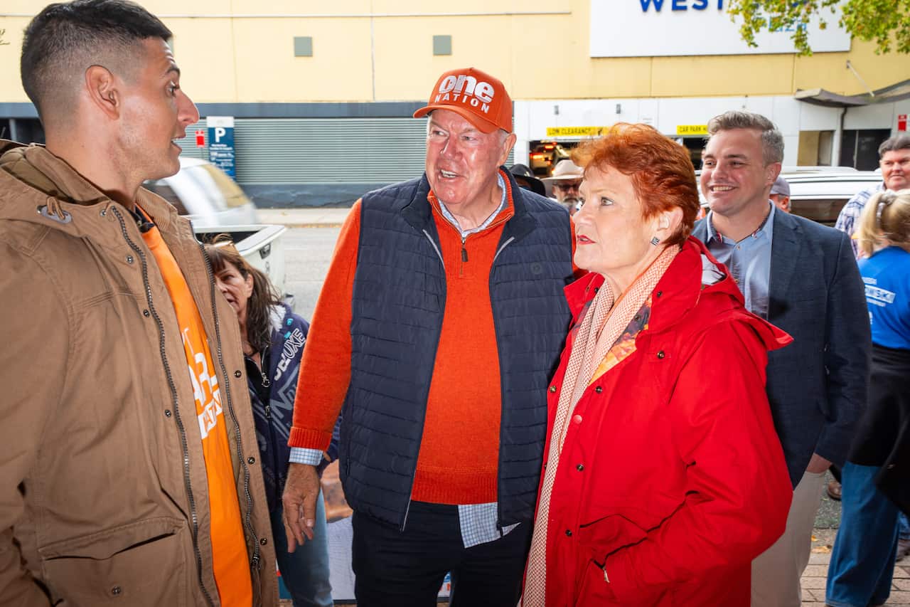 An older white man and woman wearing a red sweater and coat speak with another man 