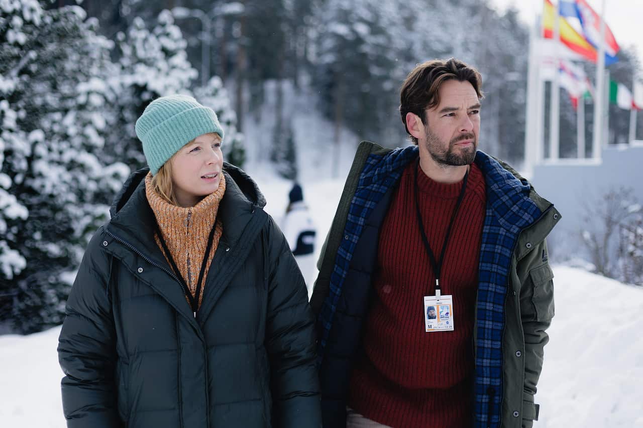 A man and a woman, wearing coats, stand outide, in the snow, with snow-covered trees behind. The corner of a structure, with national flags on poles, can be see behind them.