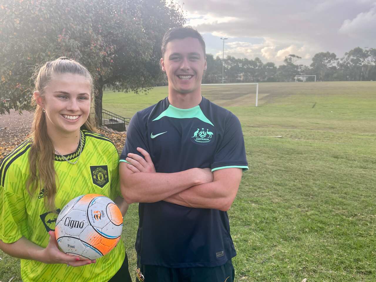 A young woman wearing a lime-coloured Manchester United football jersey and holding a ball standing next to a young man wearing a dark blue Socceroos football jersey in a park.