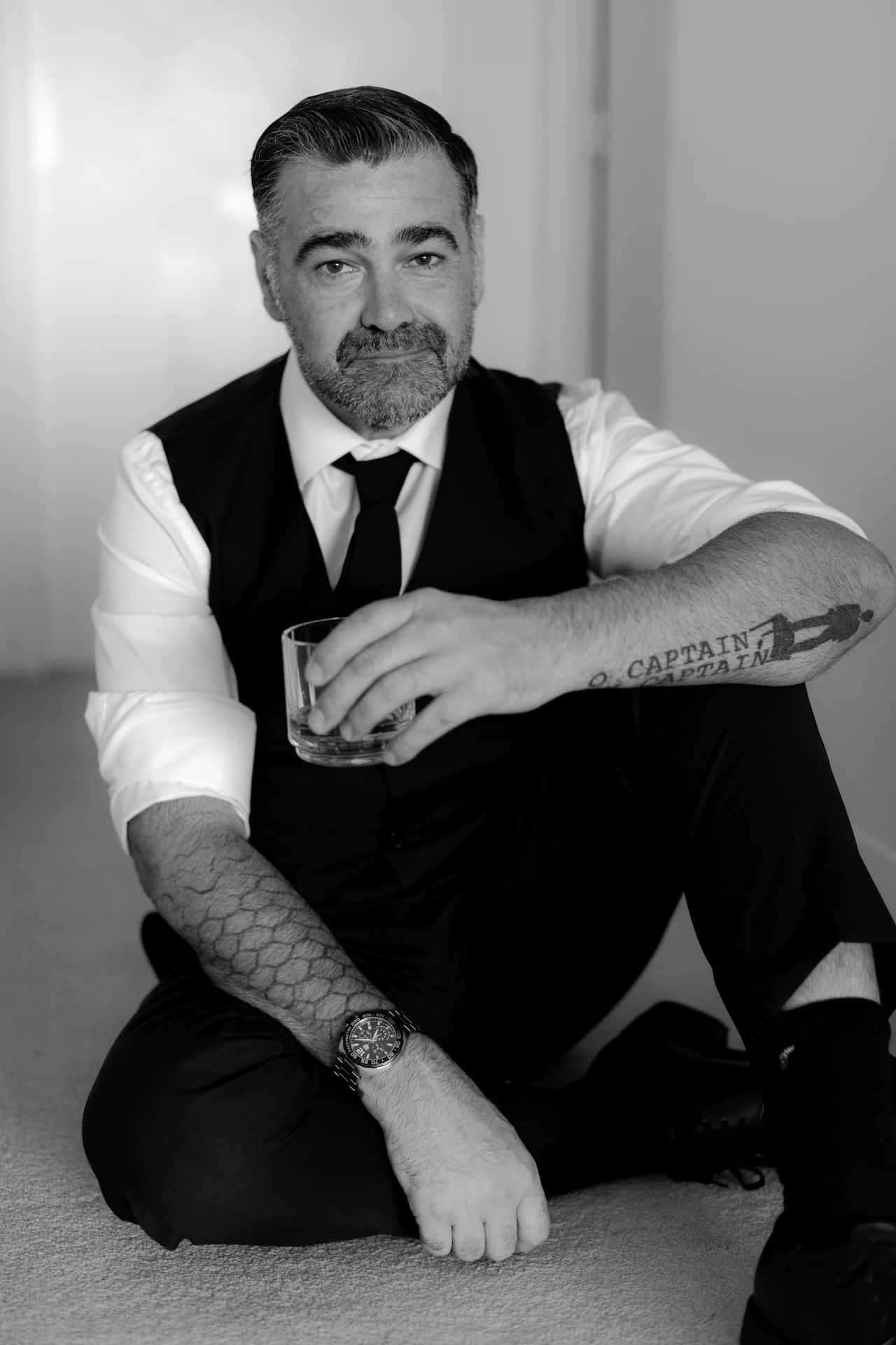 A man sitting on the ground wearing a suit and holding a glass. 