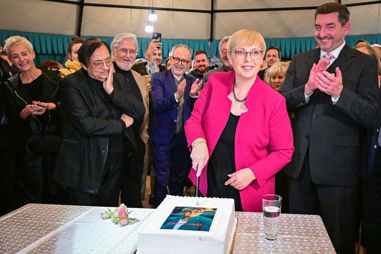 A woman in a pink jacket cuts a cake with her photo on it, surrounded by people clapping