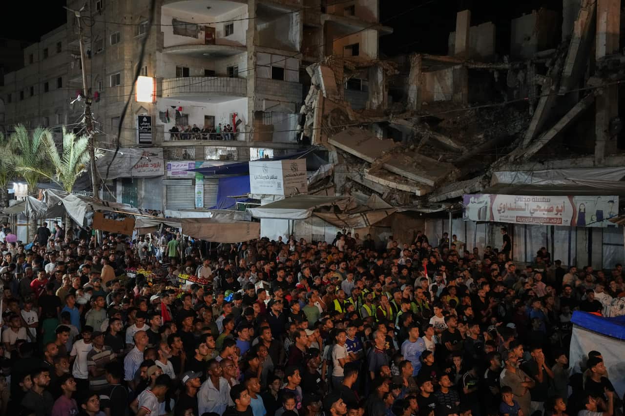 Hundreds of Palestinians crowd at the bottom of the photo all facing a screen out of shot. Behind them are the ruins of pale buildings partially collapsed.