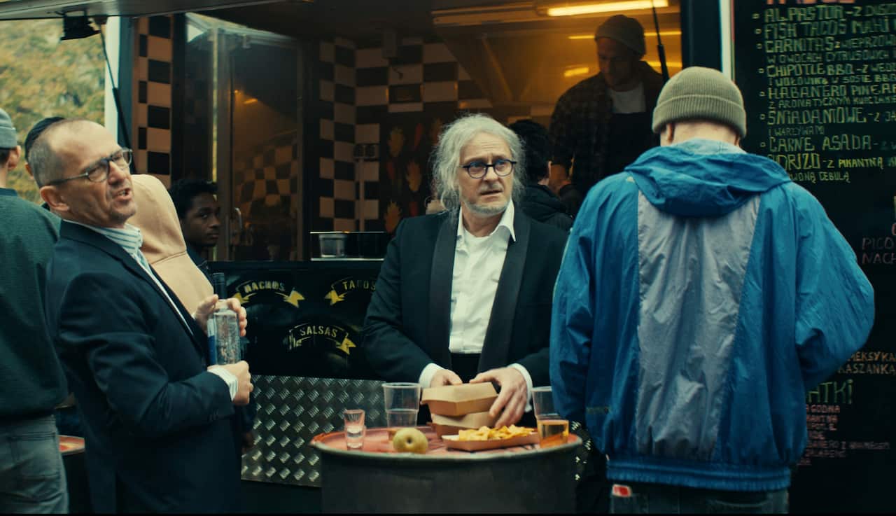 Several, including a man with white hair and glasses, stand in front of a food truck. 