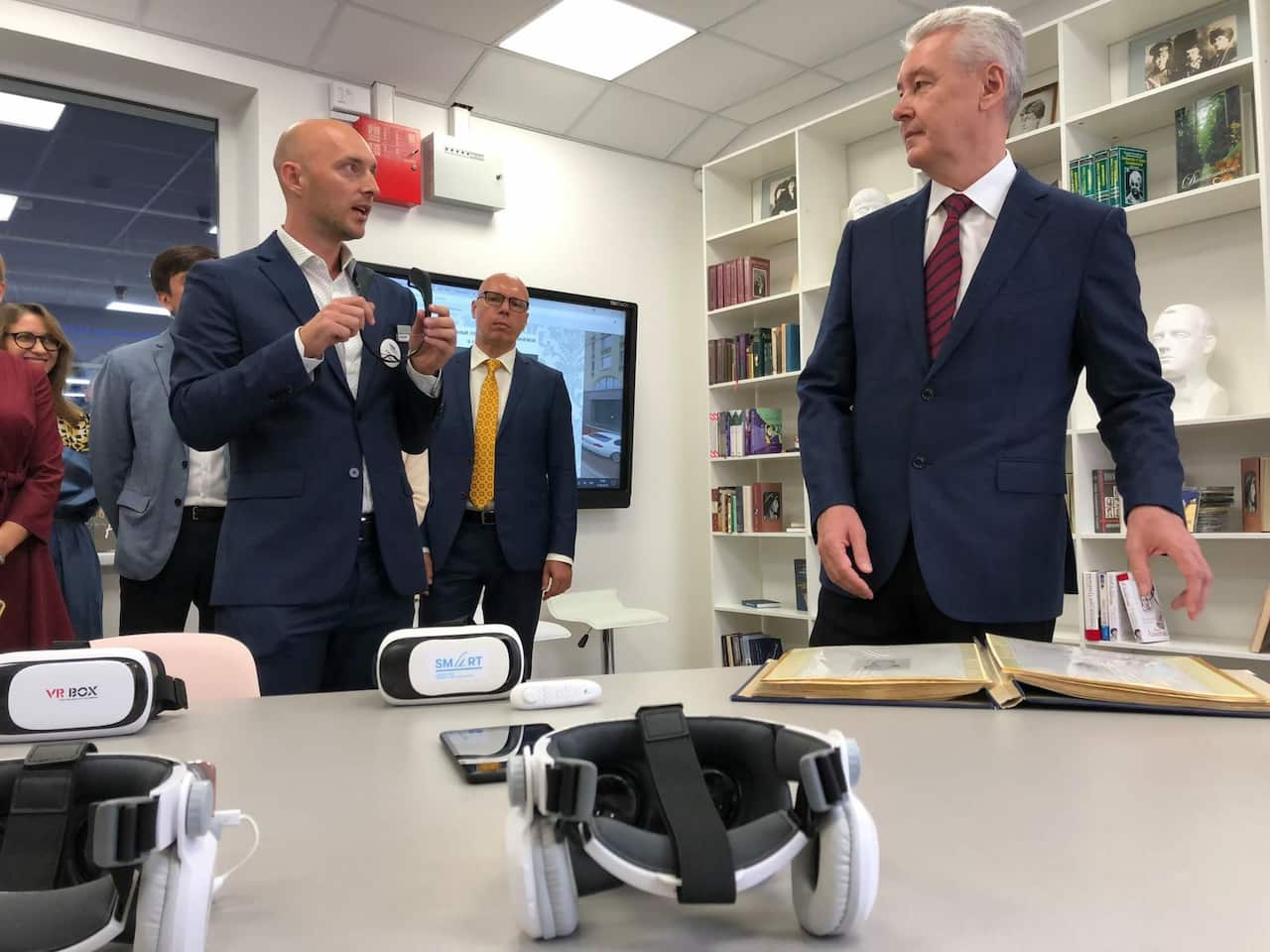 A room at a library with bookshelves and a TV screen mounted on a wall. Two men standing and talking in front of a table with VR sets. The man on the left is bald and is wearing a dark grey suit and a white shirt with an unbuttoned collar. The older man on the right has grey hair and is wearing a dark blue suit, white shirt, and a burgundy tie. 