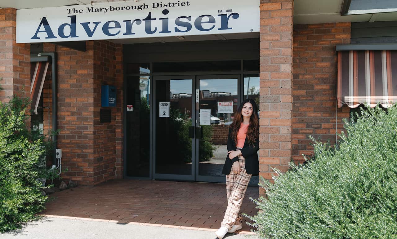 A young woman stands at entrance to a building. A sign with 'The Maryborough District Advertiser' can be seen above the doorway. 