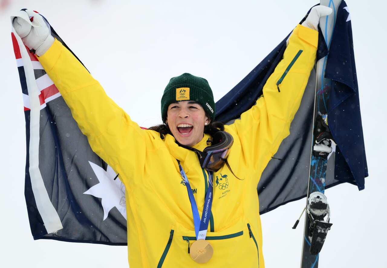 A woman wearing a yellow Australian jacket and a green beanie beams as she raises an Australian flag behind her. She's in a snowy area and clutching a ski in one hand, wearing a gold medal around her neck.