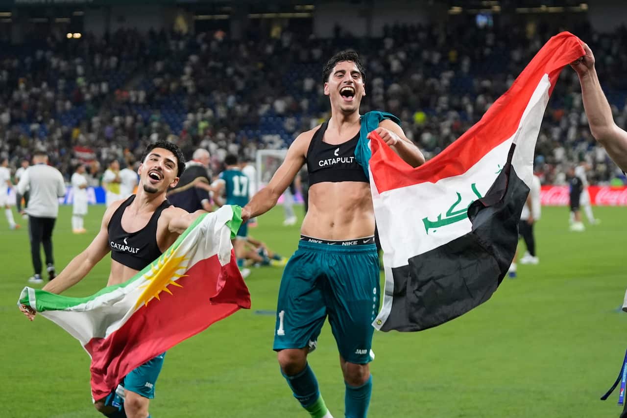Two men walk on a football field, they are sweaty and smiling, the man on the right holds the Iraqi flag. 