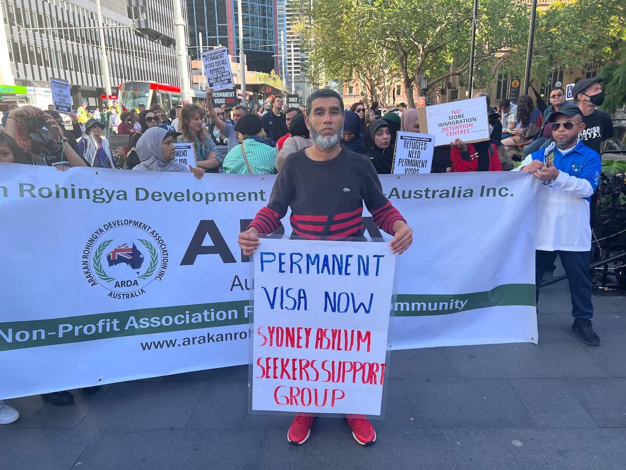  A man standing in front of people holding a banner. He holds a sign reading: 'Permanent visa now Sydney Asylum Seeker Support Group'