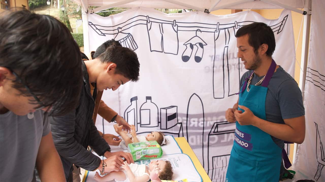 Three young men changing nappies on baby dolls with an instructor in a blue apron watching them
