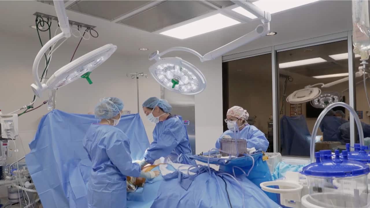 Medical staff surround an operating table during a breast reconstruction surgery. They wear light blue scrubs and the patient is covered by the same blue material.