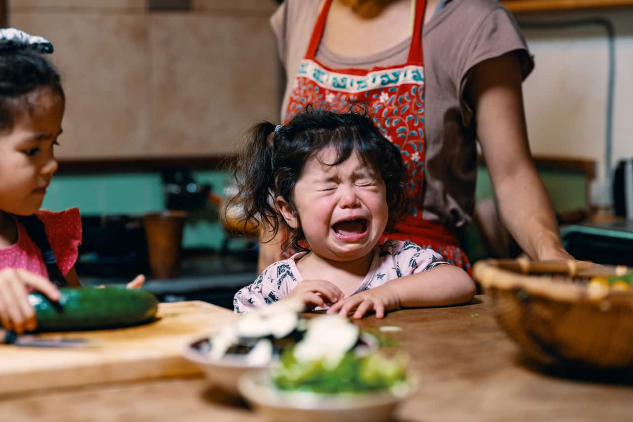 Mother preparing lunch with two young daughters as the younger one is crying