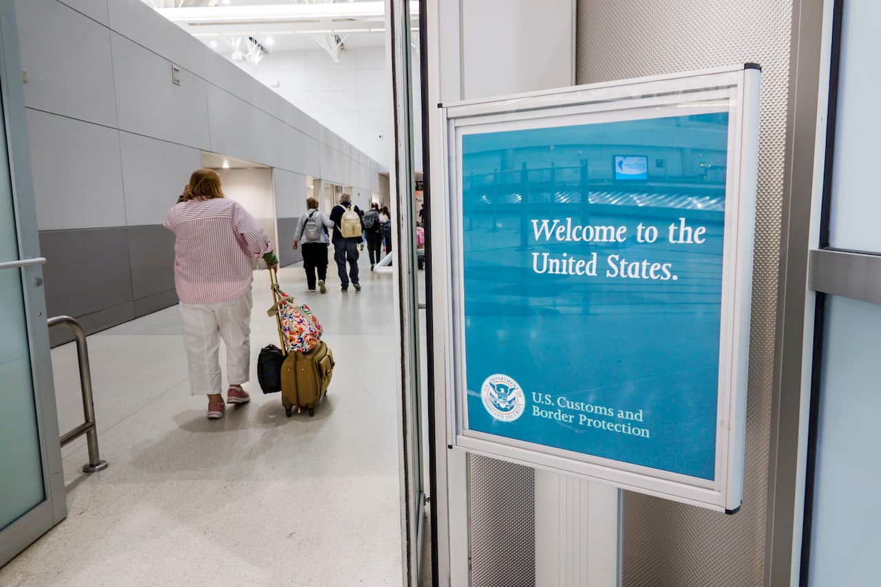 A blue sign in a metal frame next to a door at an airport terminal. The sign reads: Welcome to the United States in white print. Underneath is written: US Customs and Border protection