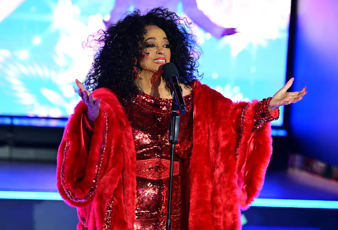 Diana Ross performs on stage, wearing a voluminous red faux-fur coat over a shimmering red sequined dress while holding a microphone.