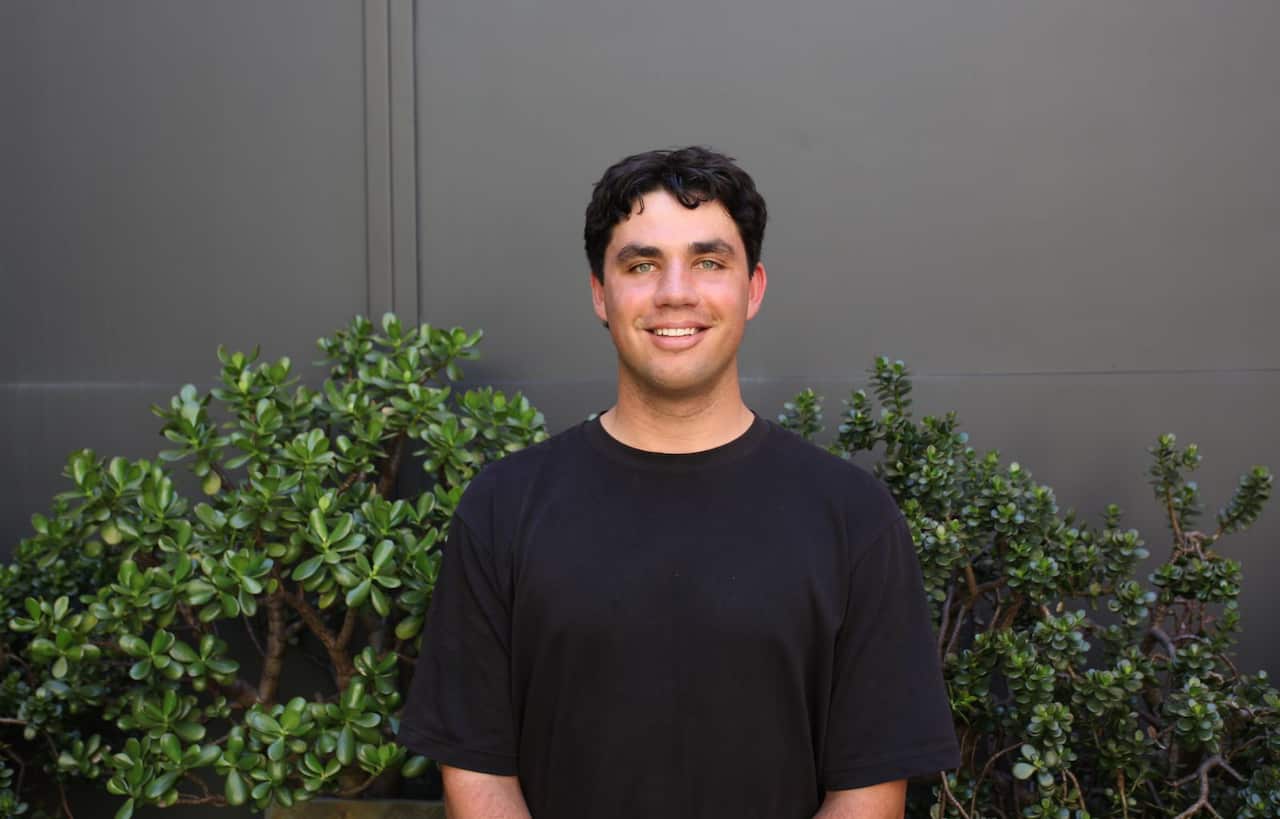 A man in a black round neck t-shirt stands in front of a bush and a wall.